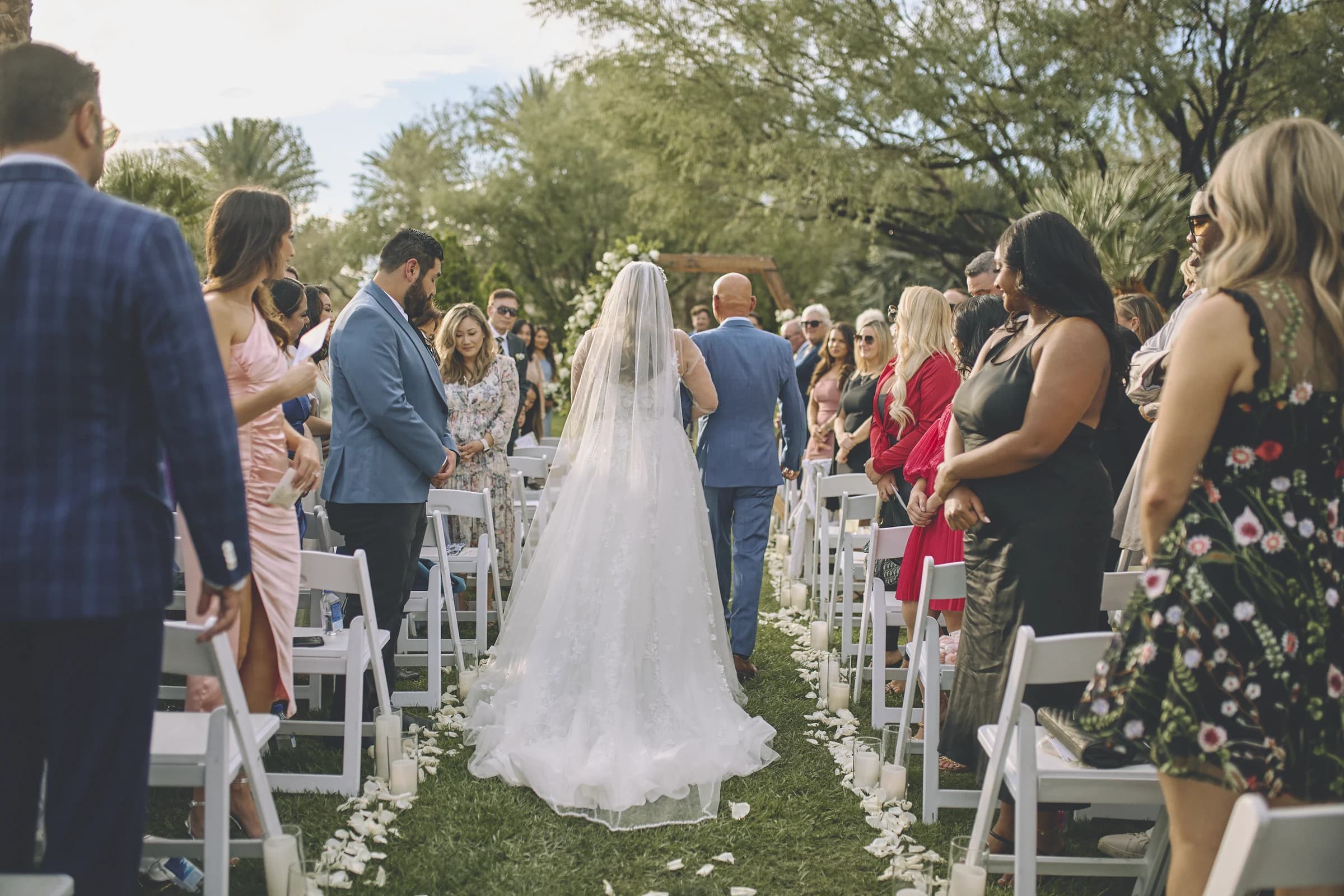 Bride and escort walk down an outdoor ceremony aisle as guests look on