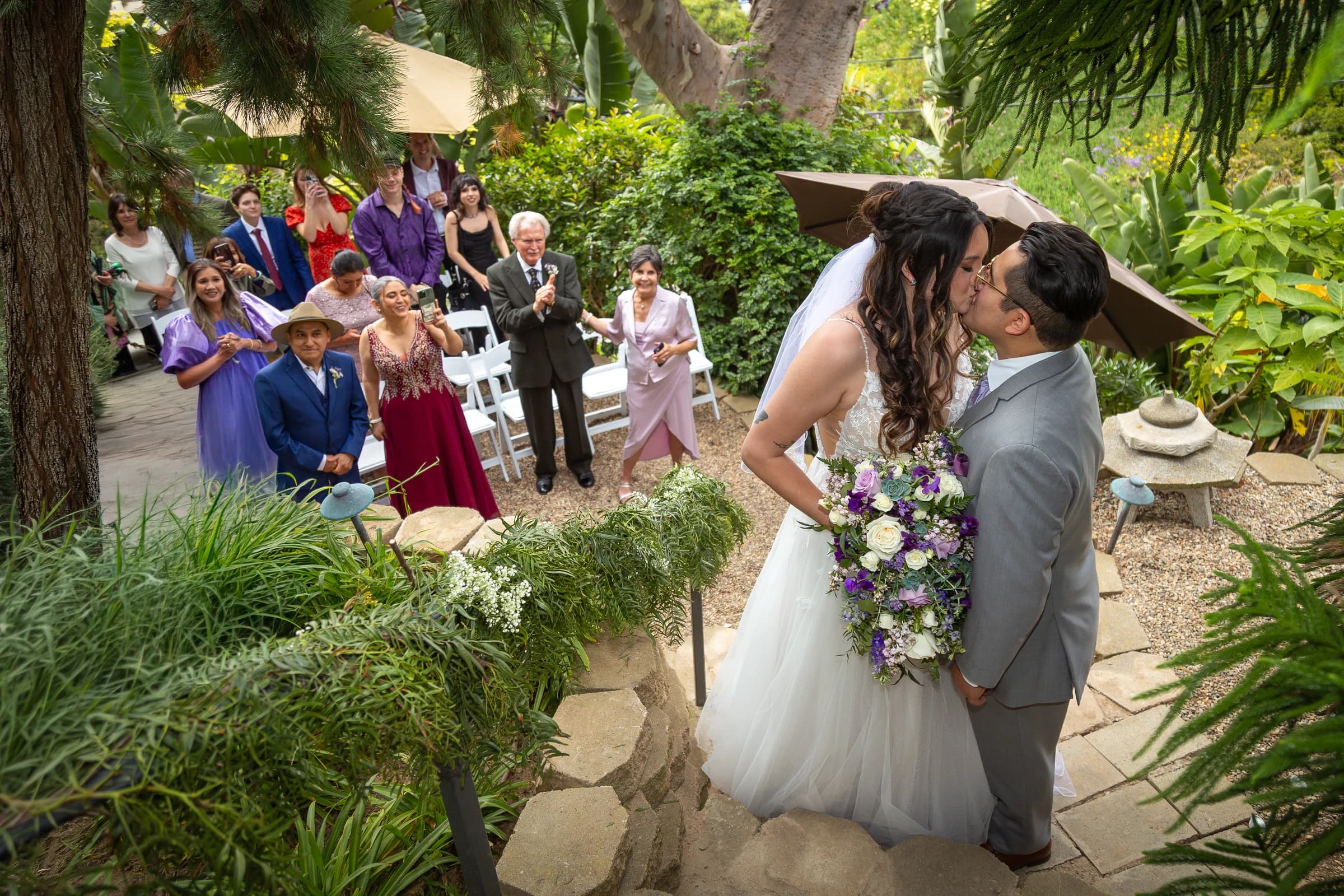 Newlyweds kiss on a garden staircase as their guests celebrate behind them