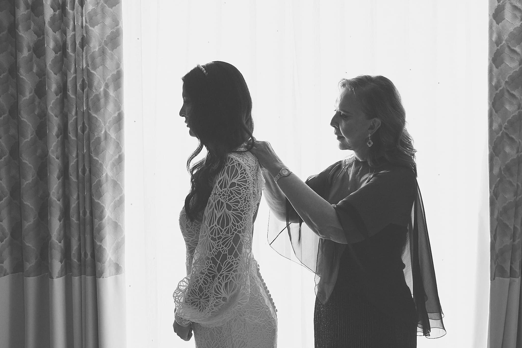 A woman adjusts a bride's veil beside a bright window in black and white